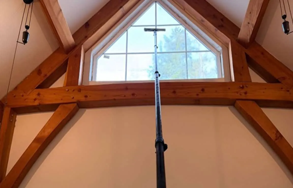 Long pole with a squeegee reaching a high triangular window framed by timber beams