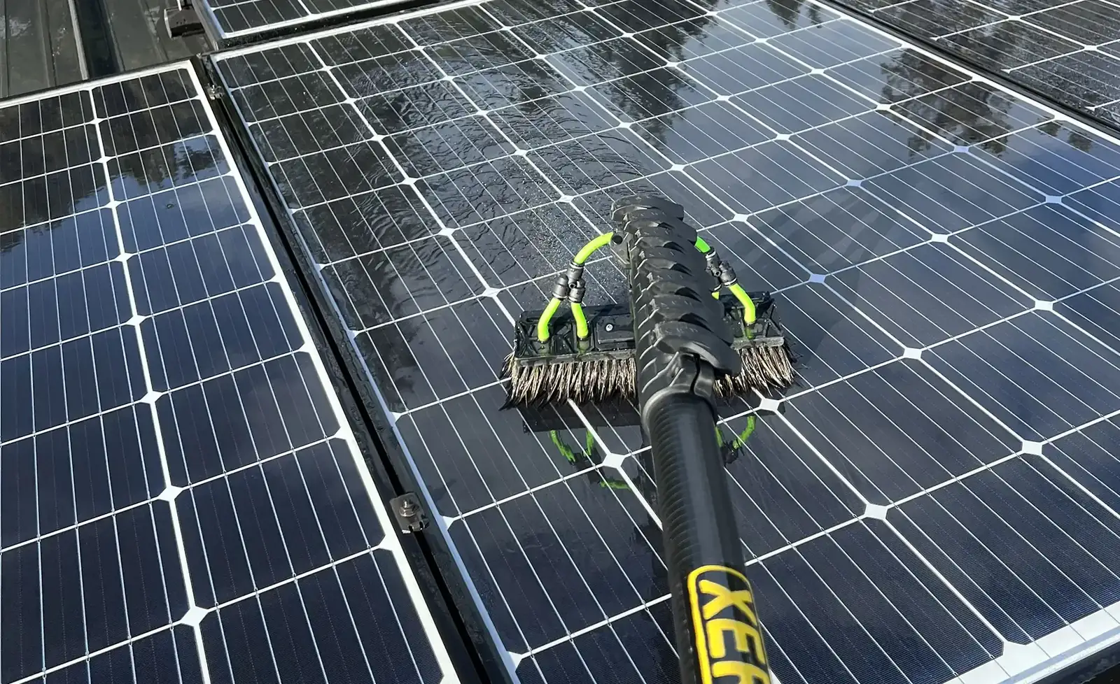 Brush on a long pole cleaning wet solar panels, water streaking across the glass