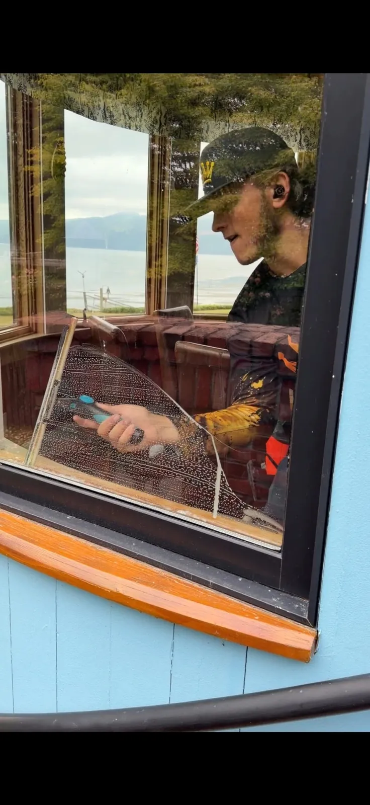 Technician using a squeegee to clean a soapy window from inside, water view beyond