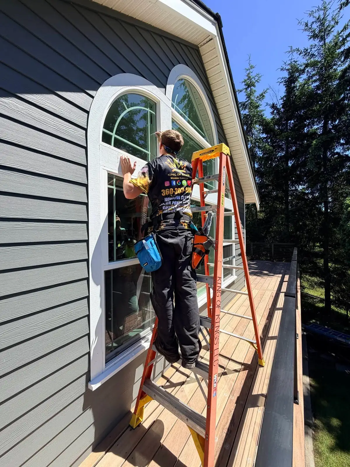 Technician on a ladder cleaning an arched second-storey window on a gray-sided house
