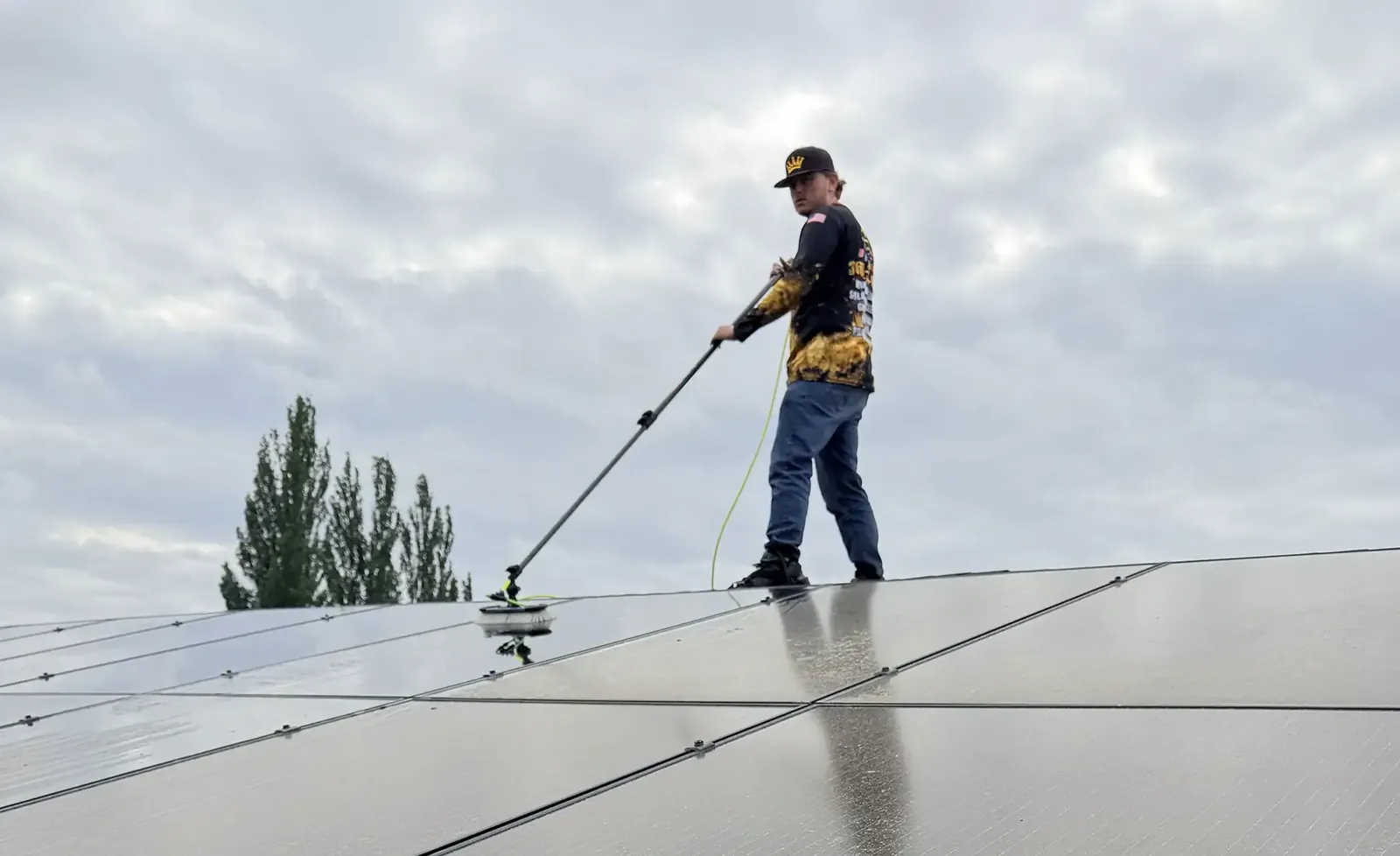 Technician standing on a rooftop cleaning solar panels with a long-handled brush